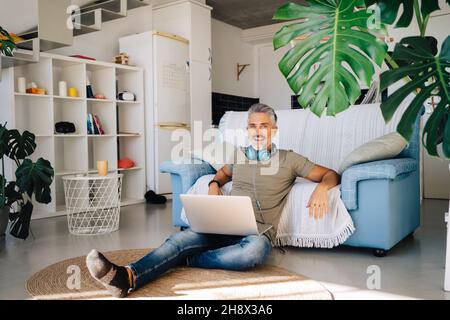 Haired grigio felice uomo bello usando il laptop e le cuffie per ascoltare la musica mentre si agghiacciano vicino a comodo divano in appartamento loft Foto Stock