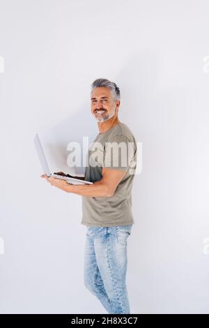 Gli anziani sorridono sorridendo il maschio guardando attentamente la macchina fotografica mentre si alza contro la parete bianca e navigando il laptop in mano nella stanza della luce Foto Stock