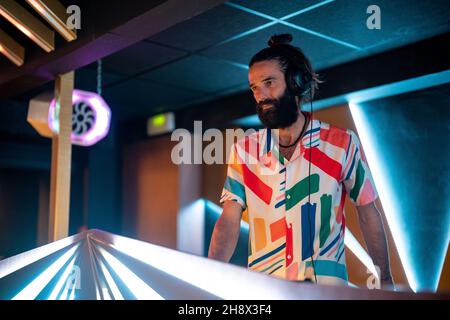 DJ maschile concentrato in cuffia in piedi al banco illuminato nel moderno nightclub Foto Stock