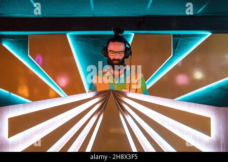 DJ maschile concentrato giovane bearded in occhiali e cuffie in piedi al banco illuminato nel moderno nightclub Foto Stock