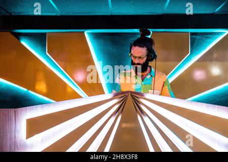 DJ maschile concentrato giovane bearded in occhiali e cuffie in piedi al banco illuminato nel moderno nightclub Foto Stock