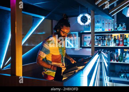 DJ maschile concentrato giovane bearded in occhiali e cuffie in piedi al banco illuminato nel moderno nightclub Foto Stock