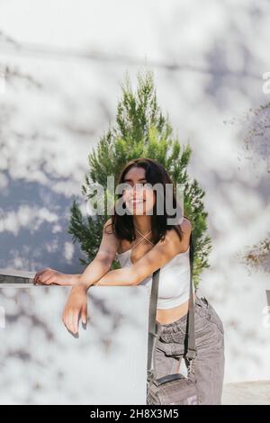 Contenuto Signora ispanica con capelli marroni appoggiati su una barriera di pietra sulla strada contro il muro e guardando via in giornata di sole Foto Stock