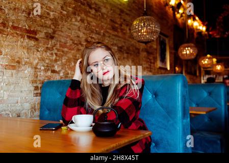 Giovane donna bionda carina e premurosa con bicchieri seduti e una tazza di tè al caffè Foto Stock