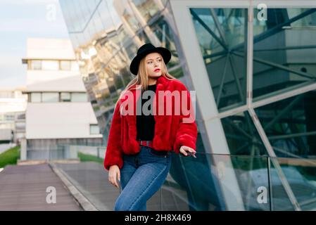 Giovane donna bionda carina con cappello e giacca rossa in piedi sulla strada e guardando via Foto Stock