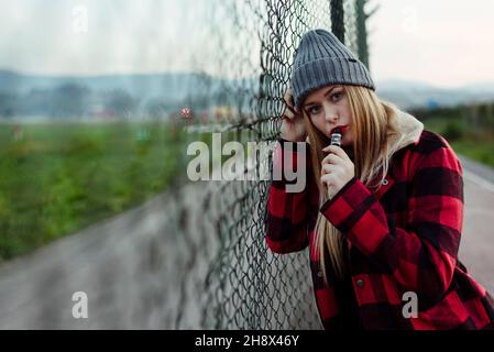 Bella giovane donna bionda con cappello di lana appoggiata sul recinto e fumare con macchina per il vapo Foto Stock