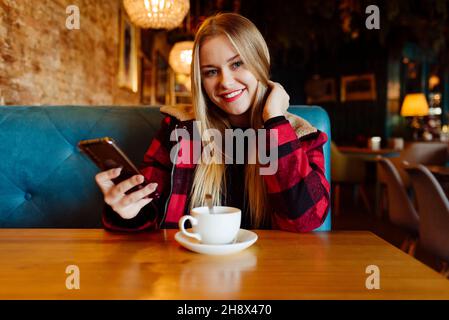 Bella giovane donna bionda che usa il telefono e che ha una tazza di tè al caffè Foto Stock