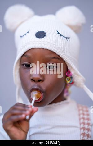 Adorabile ragazza afroamericana in caldo cappello lecca lecca lecca dolce e guardando via su sfondo grigio Foto Stock