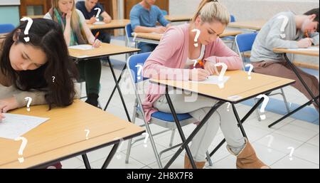 Immagine composita digitale degli studenti universitari che scrivono l'esame presso le scrivanie in aula con punti interrogativi Foto Stock