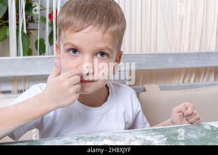 Un ragazzino emozionale e carino macchia il naso con farina e ti guarda. Gioca con la farina. Il bambino aiuta la madre con biscotti da forno, pizza a casa. Foto Stock