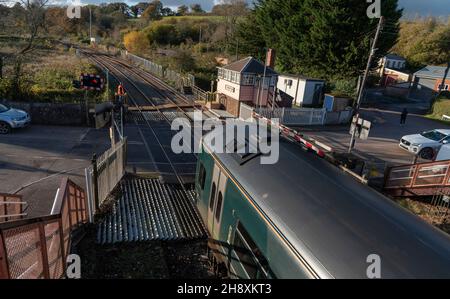 Crediton, Devon, Inghilterra, Regno Unito. 2021. Treno Passenegr con partenza dalla stazione di Crediton e passaggio a livello. Guardato da un segnalatore che emette una chiave per Th Foto Stock