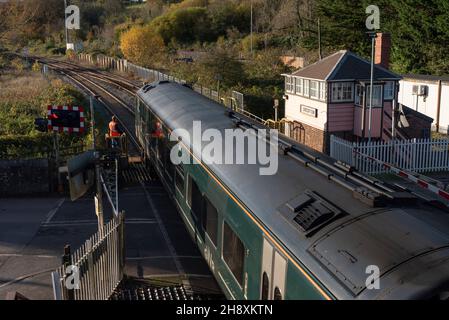 Crediton, Devon, Inghilterra, Regno Unito. 2021. Treno Passenegr con partenza dalla stazione di Crediton e passaggio a livello. Guardato da un segnalatore che emette una chiave per Th Foto Stock