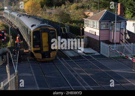 Crediton, Devon, Inghilterra, Regno Unito. 2021. Treno Passenegr con partenza dalla stazione di Crediton e passaggio a livello. Guardato da un segnalatore che emette una chiave per Th Foto Stock