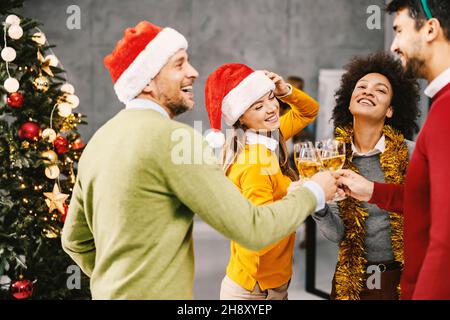 Festa di Capodanno presso l'azienda. Un gruppo di colleghi multiculturali si trova accanto a un albero di Natale e fa un brindisi con bicchieri di champagne su NE Foto Stock