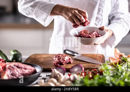 Dettaglio di pezzi di carne rossa messi in una ciotola bianca su una tavola di preparazione del pasto piena di ingedienti. Foto Stock
