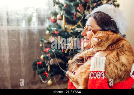 Celebrazione della Messa di Natale con cat. Donna giocando e abbracciando il pet in Santa's hat dal nuovo anno albero a casa. Foto Stock