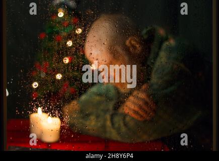 Primo piano dell'uomo anziano attento seduto accanto all'albero di Natale illuminato visto attraverso la finestra Foto Stock