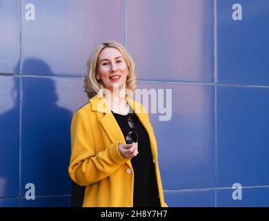Giovane donna con capelli biondi in un cappotto giallo brillante su sfondo blu su una strada della città. Stile urbano di donna moderna Foto Stock