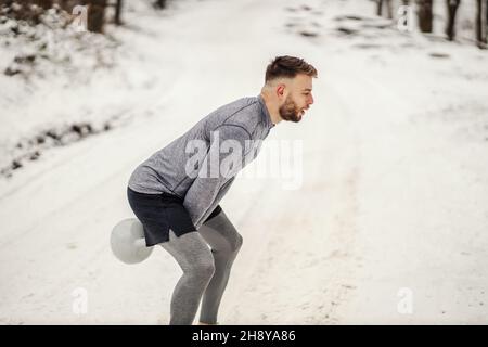 Sportivo che oscilla Kettlebell mentre in piedi in natura su sentiero innevato in inverno. Bodybuilding, sport invernali, fitness Foto Stock