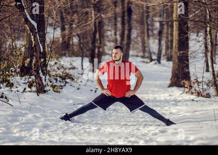 Sportivo che fa splits ed esercizi di stretching mentre si trova in natura durante la giornata invernale innevata. Esercizi di stretching, stile di vita sano, fitness invernale Foto Stock