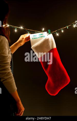 Una donna sta appendendo un avviso che dice le risoluzioni di nuovo anno ad un regalo che immagazzina su una serie di luci decorative. Un'immagine di concetto di umorismo oscuro bizzarro per un Foto Stock