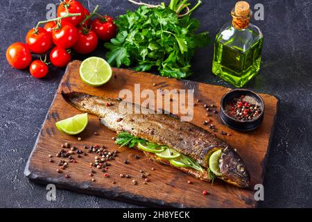 pesce intero di trota cotto in forno con erbe e spezie, servito su un tagliere di legno con fettine di lime e prezzemolo Foto Stock