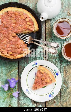 Gustosa torta di rabarbaro in padella in ghisa su tavole turchesi shabby con forchette d'epoca, tè e bluebell vista dall'alto Foto Stock