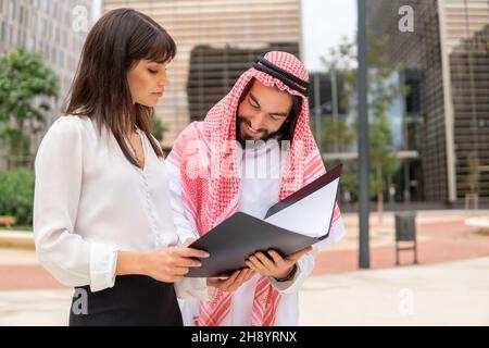 Felice uomo arabo cliente firma documenti mentre in piedi all'aperto con giovane donna d'affari caucasica, sorridente uomo d'affari arabo in abbigliamento tradizionale mettendo firma sul contratto con partner commerciale Foto Stock