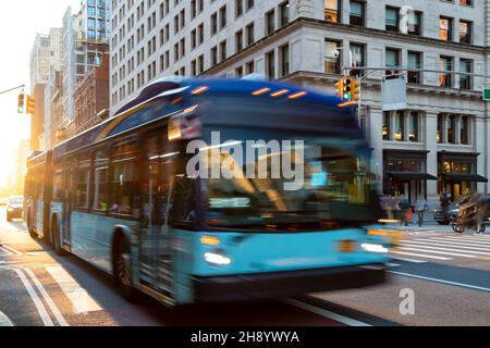 Autobus di New York City che percorrono la 23rd Street attraverso Manhattan con la luce del sole che splende tra gli edifici sullo sfondo Foto Stock