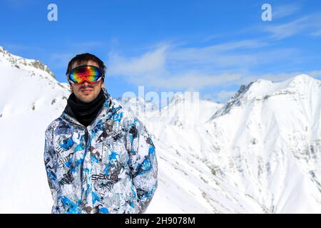 Un ragazzo in una giacca da snowboard e maschera in piedi sullo sfondo di vette innevate di montagna e cielo blu nuvoloso. Vacanze invernali attive sul monte Foto Stock