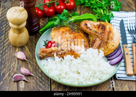 un piatto di pollo fritto alla griglia e riso bollito. pomodori, cockerel verde, aglio su un asse di legno. sfondo rustico in legno Foto Stock