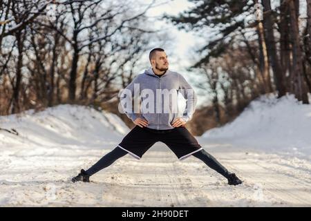 Sportivo che fa splits ed esercizi di stretching mentre si trova in natura durante la giornata invernale innevata. Esercizi di stretching, stile di vita sano, fitness invernale Foto Stock