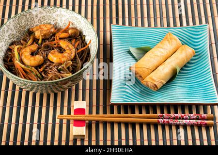 Da sopra di piatto con gustosi involtini primaverili posti vicino a ciotola con tagliatelle di yakisoba con gamberi sul tavolo Foto Stock