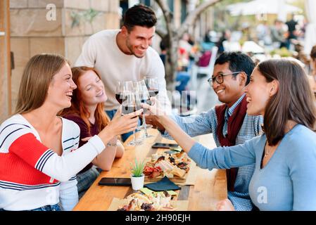 Un gruppo felice di giovani amici multiculturali che festeggiano in un ristorante, allegre e tostare con bicchieri di vino rosso mentre si siedono intorno a una lade da tavolo Foto Stock