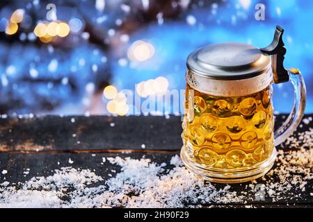 Birra di vetro stein con coperchio in metallo e rinfrescante lager servita su tavolo in legno durante la notte innevata in inverno Foto Stock