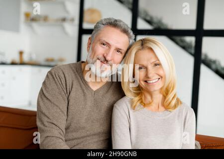 Una coppia serena e spensierata di mezza età in amore guarda la fotocamera e sorride. Bell'uomo dai capelli grigi e affascinante donna bionda si siedono in abbraccio sul divano. Concetto di amore e legame Foto Stock