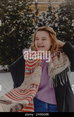 Giovane donna bionda in sciarpa con motivi ride sullo sfondo di alberi di natale nella neve. Stagione invernale. Foto Stock