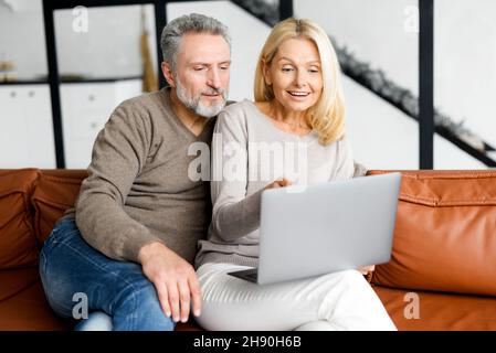 Coppia allegra di mezza età con un computer portatile seduto sul comodo divano in pelle a casa, scegliendo nuovi mobili, facendo ordine di cibo, scorrimento news feed. I coniugi maturi trascorrono del tempo libero insieme Foto Stock