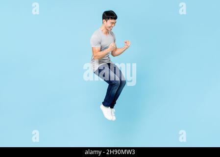 Ritratto di sorridente giovane uomo asiatico felice che aggrappa pugni e salta su isolato sfondo azzurro studio Foto Stock