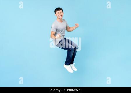 Ritratto di sorridente giovane uomo asiatico felice che aggrappa pugni e salta su isolato sfondo azzurro studio Foto Stock