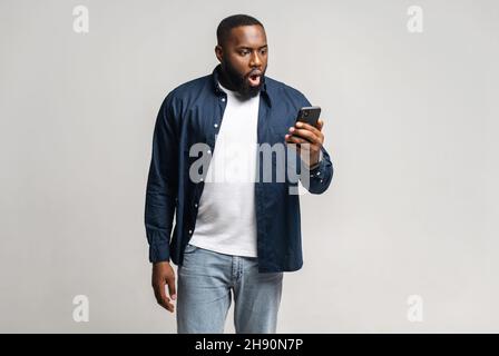 Scioccato e sguardato afroamericano che tiene smartphone e guarda lo schermo, ritratto di nero pop-eyed uomo e aperto bocca guardando con incredulo al telefono display isolato sul grigio Foto Stock