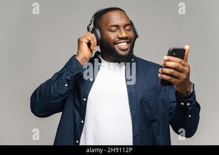 L'esilarante uomo afro-americano che indossa le cuffie utilizzando uno smartphone, spende il tempo libero online, guardando video, ascoltando musica e sorrisi, felice ragazzo nero con le cuffie tiene il telefono, isolato su gary Foto Stock