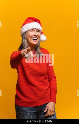 Allegro felice donna anziana in santa Hat punti indice dito a macchina fotografica e ride, verticale ritratto di mezza età femmina scelta voi. Nuovo anno di pubblicità concetto Foto Stock