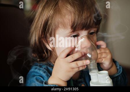 Bambina in maschera, cura il tratto respiratorio con un nebulizzatore a casa. Il bambino siede con un nebulizzatore nella sua bocca, inalatore, trattamento della bronchite Foto Stock