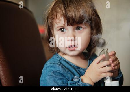 Bambina in maschera, cura il tratto respiratorio con un nebulizzatore a casa. Il bambino siede con un nebulizzatore nella sua bocca, inalatore, trattamento della bronchite Foto Stock