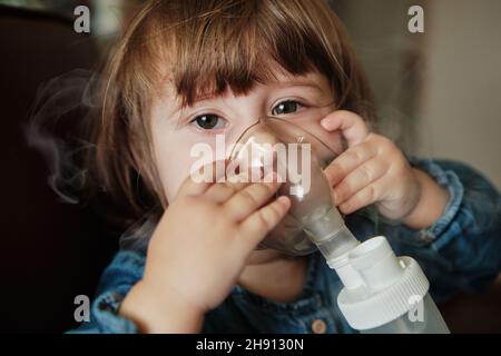 Bambina in maschera, cura il tratto respiratorio con un nebulizzatore a casa. Il bambino siede con un nebulizzatore nella sua bocca, inalatore, trattamento della bronchite Foto Stock