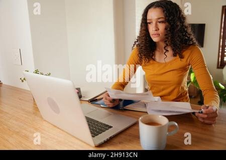 Donna multiculturale interessata, pagare le fatture. Giovani professionisti che lavorano a distanza con il computer portatile in un appartamento moderno Foto Stock