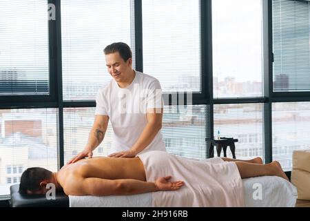 Vista laterale del massaggiatore professionale maschile con mani tatuate e forti che massaggiano schiena e spalle dell'uomo sportivo muscolare sdraiato sullo stomaco Foto Stock