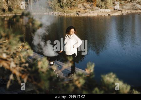 Donna incinta sorridente che prepara il cibo al lago Foto Stock