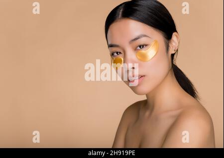 Cerotti oculari in oro per puffiness, rughe e cerchi scuri, concetto di cura della pelle. Bella giovane donna bruna asiatica con pelle perfetta applicata gold collagene patch occhio, in piedi su isolato sfondo beige Foto Stock